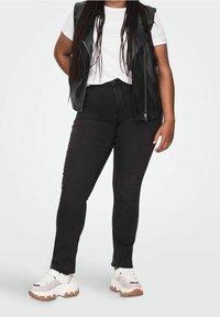 Černá kožená vesta přes bílý tričko, doplněná o černé skinny džíny a robustní bílé tenisky s hnědými akcenty.