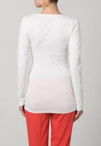 Witte langemouwen top met een bootnek, gemaakt van zachte stof. De achterkant is glad en aansluitend, in contrast met helderrode broek.