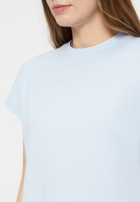 Femme aux cheveux châtain clair portant un t-shirt uni bleu clair à col rond, vue des épaules aux lèvres sur fond blanc.