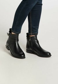 Botine negre din piele cu vârf ascuțit, închidere cu fermoar și curele decorative. Textură netedă și talpă plată. Purtați cu blugi închiși la culoare.