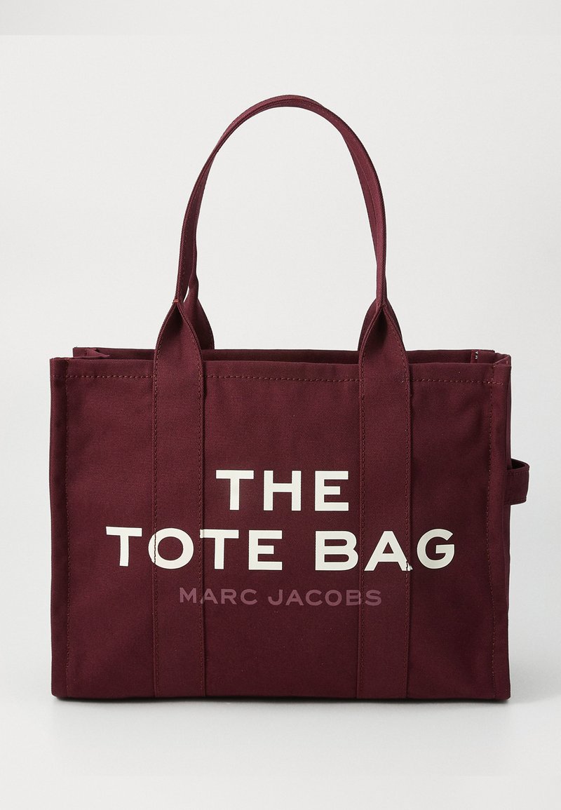 Burgundowa torba wykonana z płótna z wzmocnionymi uchwytami, z białym napisem "THE TOTE BAG" i mniejszym napisem "MARC JACOBS" na przedniej stronie.