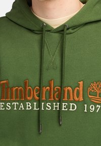 Felpa verde con cappuccio caratterizzata da un logo "Timberland" ricamato in arancione e bianco. Include lacci con punte in argento e dettagli a coste.