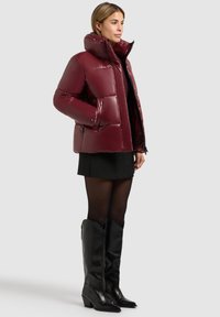 Bordeauxfarbene Pufferjacke mit hohem Kragen, glänzendem Material, großen, gesteppten Abschnitten und zwei seitlichen Taschen, kombiniert mit einem schwarzen Rock und kniehohen Stiefeln.