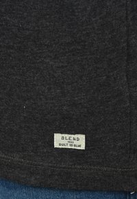 Tela de camiseta gris oscuro con una textura suave. Presenta una pequeña etiqueta blanca con "BLEND 1993 BUILT IN BLUE" cosida en la parte inferior.