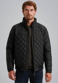 Zwarte gewatteerde jas met een staande kraag, ritssluiting aan de voorkant en zichtbare stikseldetails. Bevat ribbels bij de manchetten en heeft een iets cropped pasvorm.