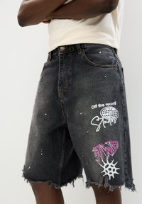 Pantaloni shorts in denim nero con orli sfrangiati, caratterizzati da macchie di vernice bianca e stampe grafiche colorate in viola e bianco sul davanti.