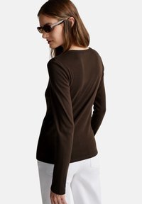 Długi rękaw, dopasowana bluzka w ciemnobrązowym materiale. Posiada gładką teksturę i klasyczny okrągły dekolt, zestawiona z białymi spodniami.