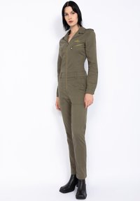 Tuta verde oliva con chiusura zip sul davanti, due tasche sul petto e passanti per cintura. Caratterizzata da maniche lunghe, colletto e design aderente. Stivaletti neri alla caviglia.