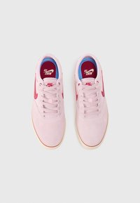 Sapatilhas de camurça rosa claro com detalhes em vermelho e atacadores, apresentando uma palmilha azul. O design inclui um logotipo swoosh visível de cada lado.
