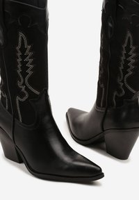 Bottes de cowboy noires avec un orteil pointu et un talon carré. Fabriquées en cuir avec des accents texturés et des coutures blanches contrastantes sur la tige.