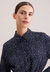 Camicia blu navy con bottoni a vista, caratterizzata da pois bianchi, colletto a punta e una texture di tessuto liscia. Progettata con una vestibilità standard e bottoni classici.