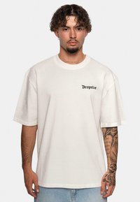 Tricou din bumbac de mari dimensiuni, de culoare crem, având un logo negru "Dropsize" pe partea stângă a pieptului. Mâneci scurte, guler rotund, croială lejeră.