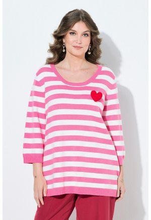 Kvinde med bølget hår iført en lyserød og hvidstribet sweater med rødt hjerte og røde bukser, stående foran en ensfarvet hvid baggrund.