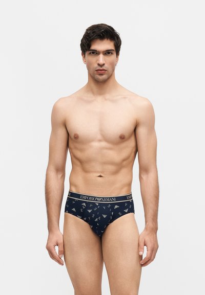 Αθλητικός άνδρας που στέκεται και κοιτάζει μπροστά, φορώντας σκούρο μπλε σλιπ Emporio Armani με μοτίβο λογότυπου σε απλό λευκό φόντο.