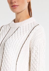 Weißer Strickpullover mit strukturiertem Muster, geripptem Ausschnitt und goldenen Kettenakzenten entlang der Nähte für zusätzliche Details.