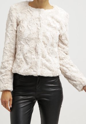 Mujer con una chaqueta blanca texturizada de pelo sintético y pantalones de cuero negro sobre un fondo claro y sencillo.