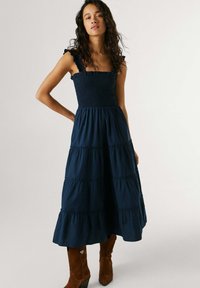 Ženska oblečena v temno modro midi obleko brez rokavov z nabranimi trakovi in rjavimi škornji do kolen stoji z eno roko na boku.