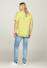 Koszulka z krótkim rękawem w kolorze limonkowym, o luźnym kroju, z lekkiego materiału, z zaokrąglonym dołem i lekkimi rozcięciami po bokach, zestawiona z jasnoniebieskimi dżinsami i białymi trampkami.