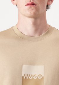Beige kokvilnas T-krekls ar apaļu kaklu. Iekļauts kvadrātveida uzlikums ar uzrakstu "HUGO" baltajā krāsā. Gluda tekstūra, minimāls dizains.