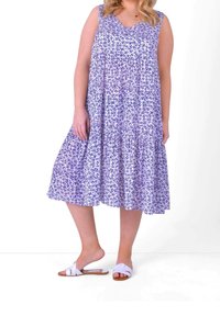 Woman wearing a sleeveless purple floral midi dress and white flat sandals, standing with one foot slightly raised.