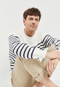 Maglione a righe blu navy e bianche con scollatura a barca e tre bottoni sulla spalla, abbinato a pantaloni beige arrotolati.