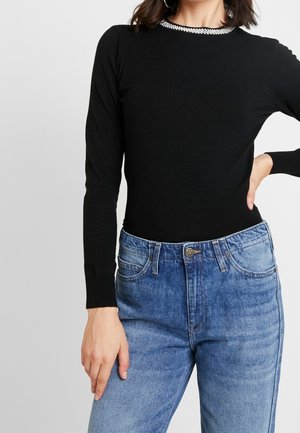 Frau trägt einen schwarzen Langarmpullover mit einem Strasskragen und hoch taillierten blauen Jeans, Hand auf der Hüfte ruhend.