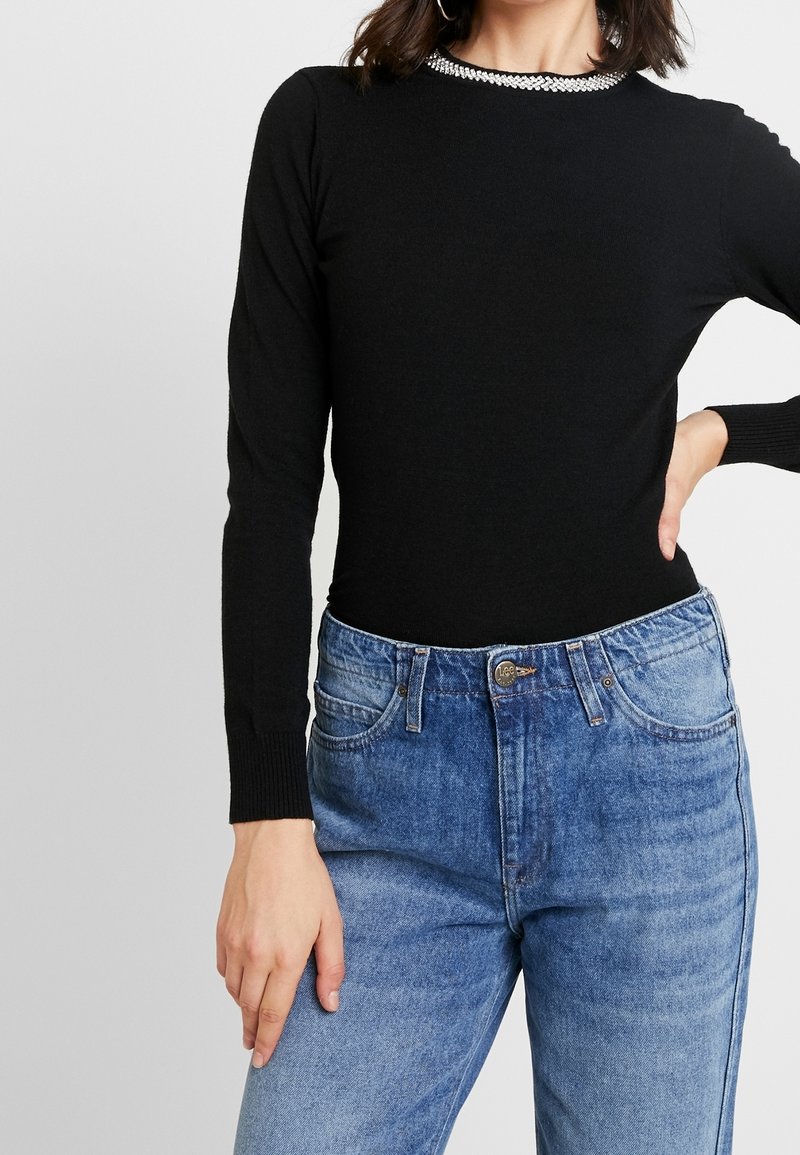 Femme portant un pull noir à manches longues avec un col orné de strass et un jean bleu taille haute, la main posée sur la hanche.
