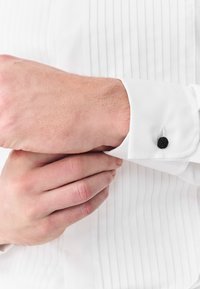 Valkoinen kauluspaita, jossa on laskostettu muotoilu. Kalvosin kiinnitetty mustalla, teksturoidulla solmu-napilla. Käsi säätää kalvosinta.