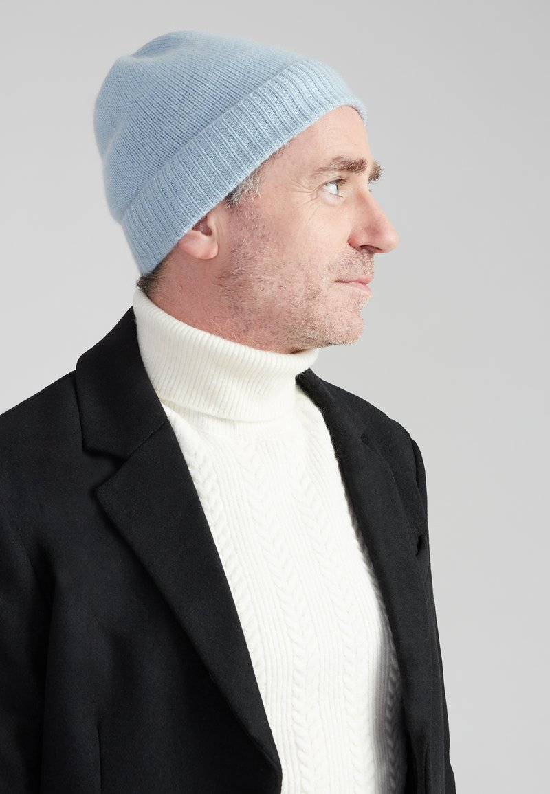 Lichtblauwe gebreide beanie met geribbelde rand, gedragen met een crèmekleurige coltrui en een zwarte blazer. Close-up profielweergave.