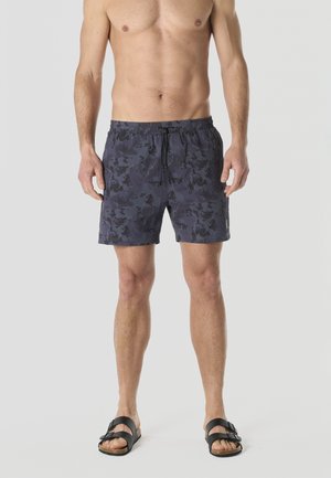 Hombre sin camisa, con pantalones cortos de baño azul oscuro con estampado y sandalias negras de dos tiras, de pie contra un fondo liso.