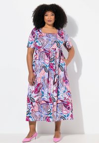 Vestido largo floral en un patrón de paisley en púrpura, rosa y azul. Mangas cortas, escote cuadrado, con costura en la cintura y bolsillos. Combinado con tacones rosas.
