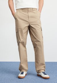 Beige cargo pants with multiple pockets and a straight cut. Worn with white sneakers featuring blue laces and brown soles on a blue surface.