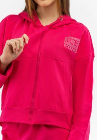 Sudadera con cremallera de color fucsia con capucha y bolsillos frontales. Presenta un logotipo de strass en el pecho y puños y cinturilla acanalados. Tejido suave y texturizado.