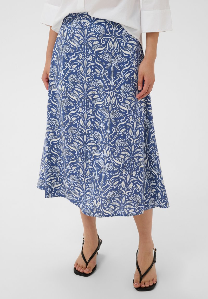 Jupe midi à motif floral bleu et blanc portée avec un chemisier blanc et des sandales noires à brides sur une personne debout sur fond blanc.
