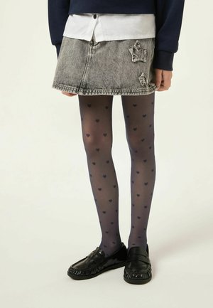 Personne portant une jupe en jean grise délavée, des collants bleu marine à motifs de cœurs, des mocassins noirs, une chemise blanche et un pull bleu marine, debout.