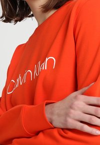 Světlý oranžový svetr s bílým logem "Calvin Klein". Má žebrované manžety a kulatý výstřih; hladká textura a ležérní střih.