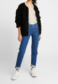 Veste bomber en velours noir, pull crème à côtes, jean bleu taille haute, baskets blanches, manches retroussées et une ceinture blanche.