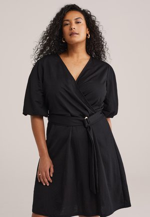 Kohvris lokkis juustega naine, kes kannab musta vöökinnitusega kleiti, millel on volangvarrukad ja vöö ümber talje, seisab ühevärvilise tausta ees.