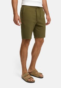 Hombre vestido con pantalones cortos verde oliva con cordón y sandalias de ante color beige, de pie frente a un fondo blanco.