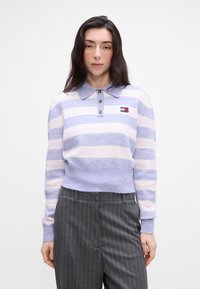 Pulover tricotat în dungi lila și roz pal, cu guler, trei nasturi și un logo mic. Purtați cu pantaloni gri cu dungi, croială scurtă.