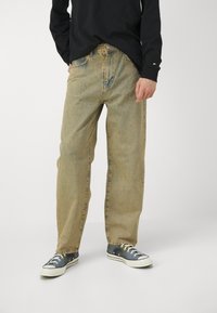 Light wash denim pants with a relaxed fit, featuring a button and zipper fly, five pockets, and subtle fading details. Paired with dark sneakers.