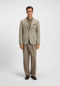 Hellgrauer strukturierter Blazer mit zwei Taschen, khakifarbener Poloshirt und beigen weit geschnittenen Hosen. Braune Schuhe runden den formellen Look ab.
