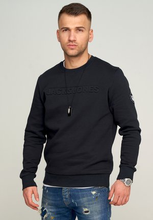 Black sweatshirt with embossed "JACK & JONES" logo. Features a crew neck, long sleeves, and ribbed cuffs. Worn with distressed blue jeans.