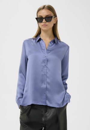 Blusa di satin azzurro chiaro con colletto, chiusura frontale con bottoni e maniche lunghe; abbinata a pantaloni in pelle neri.