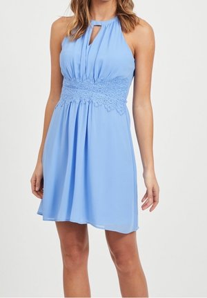 Ærmeløs lyseblå kjole med halterneck, nøglehuldsdetalje og bredt blonderbånd ved taljen, der falder over knæene på en stående kvinde.