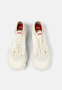 Sneaker bianche con tomaia in rete e dettagli lisci. Presentano lacci, etichette rosse con logo e una suola spessa e texturizzata.