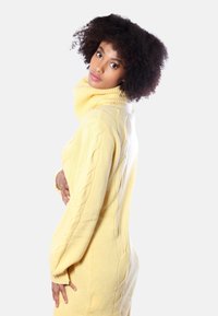 Maglione giallo lavorato a maglia con colletto alto a coste, decorato con motivi a treccia sulle maniche e sui lati, texture morbida e vestibilità aderente.