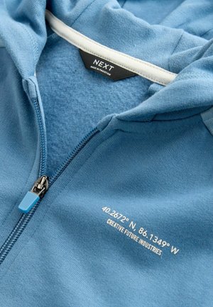 Blå hættetrøje med lynlås lavet af blødt stof. Har en hvid inderforing, en blå lynlåstrækker og hvid tekst med geografiske koordinater og mærkenavn.