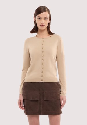 Donna che indossa un cardigan beige con bottoni e una gonna di suede marrone scuro con grandi tasche frontali, in piedi davanti a uno sfondo chiaro e semplice.