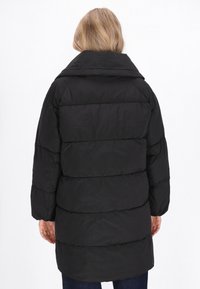 Fekete puffertakaró magas gallérral, steppelt kivitelben, hosszú ujjakkal. Az anyag simának tűnik, enyhén fényes felülettel.